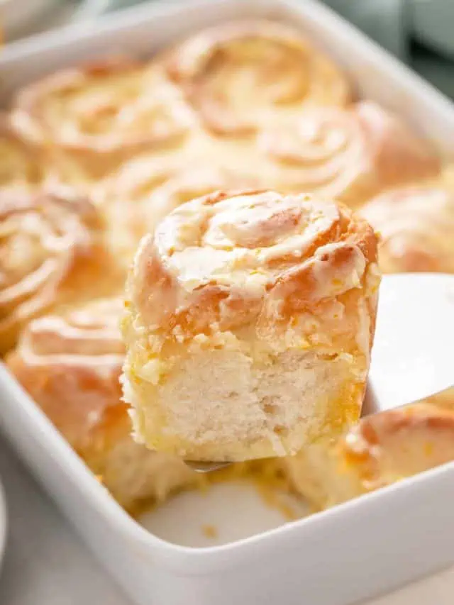 Cake server lifting up a frosted orange roll from the baking dish.