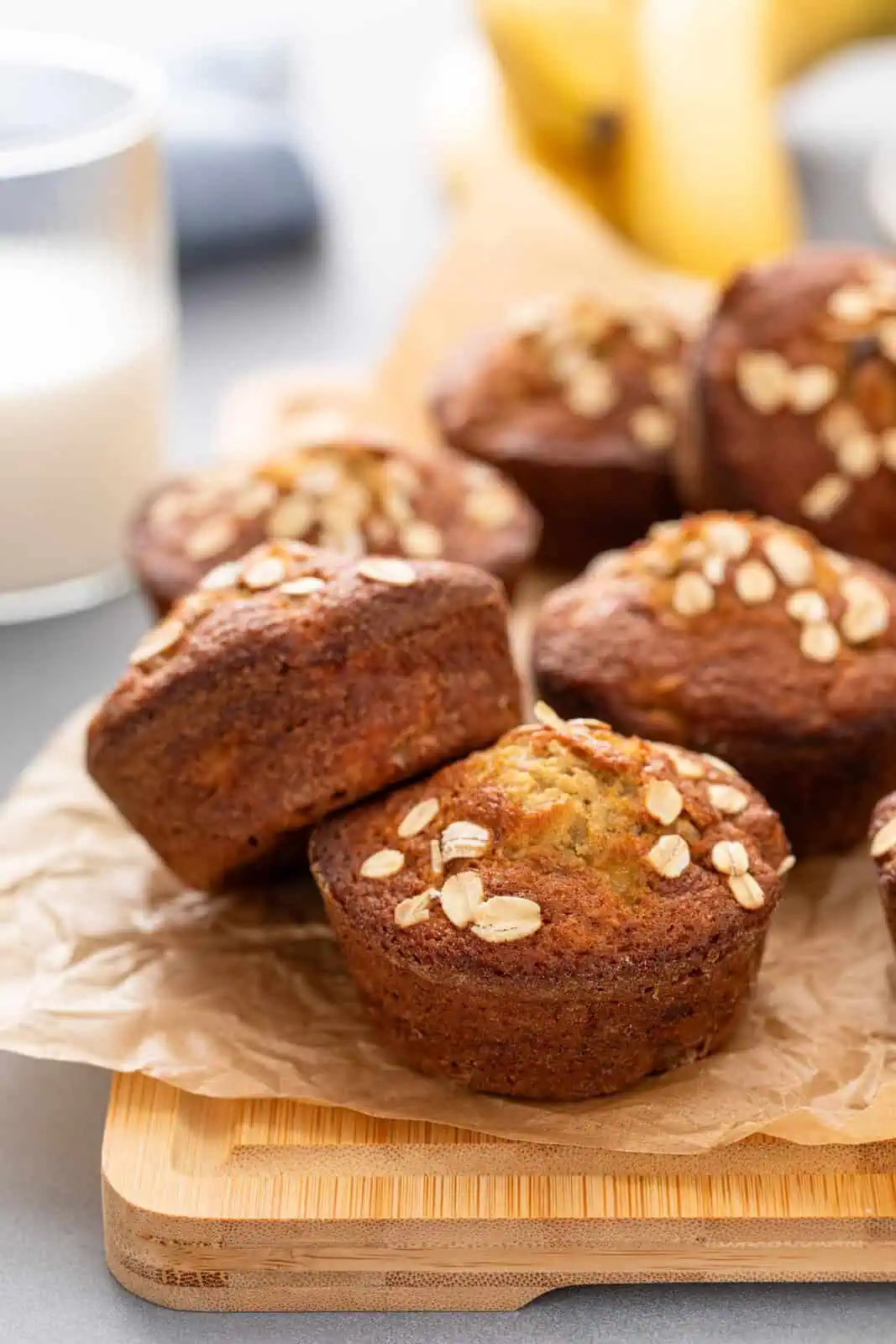 Banana oat muffins topped with rolled oats on a board in front of a glass of milk.