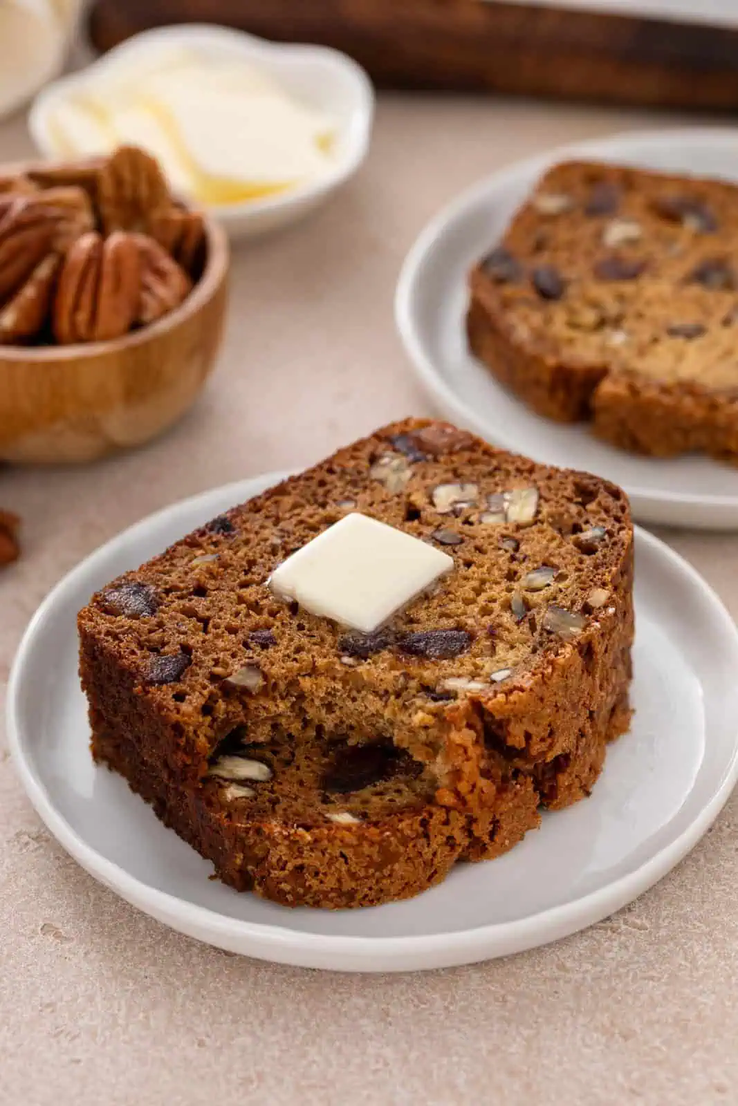 Two slices of date nut bread stacked on a white plate. A pat of butter is on the top slice, which has a bite taken from the corner.