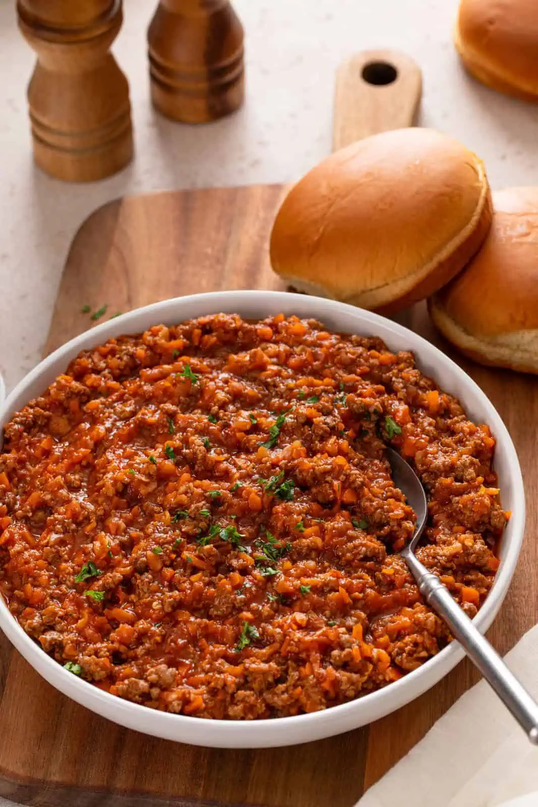 Shallow white bowl filled with sloppy joe meat on a wooden board next to hamburger buns.