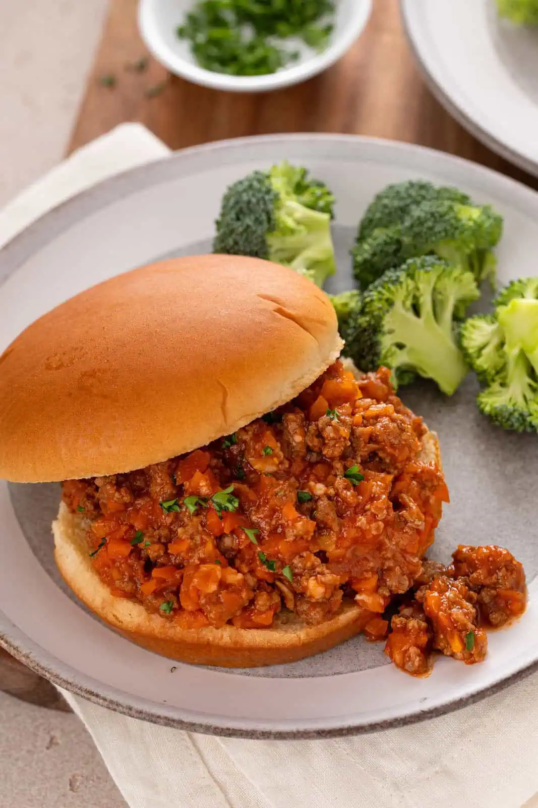 Close up image of a sloppy joe sandwich on a bun set on a plate next to broccoli.