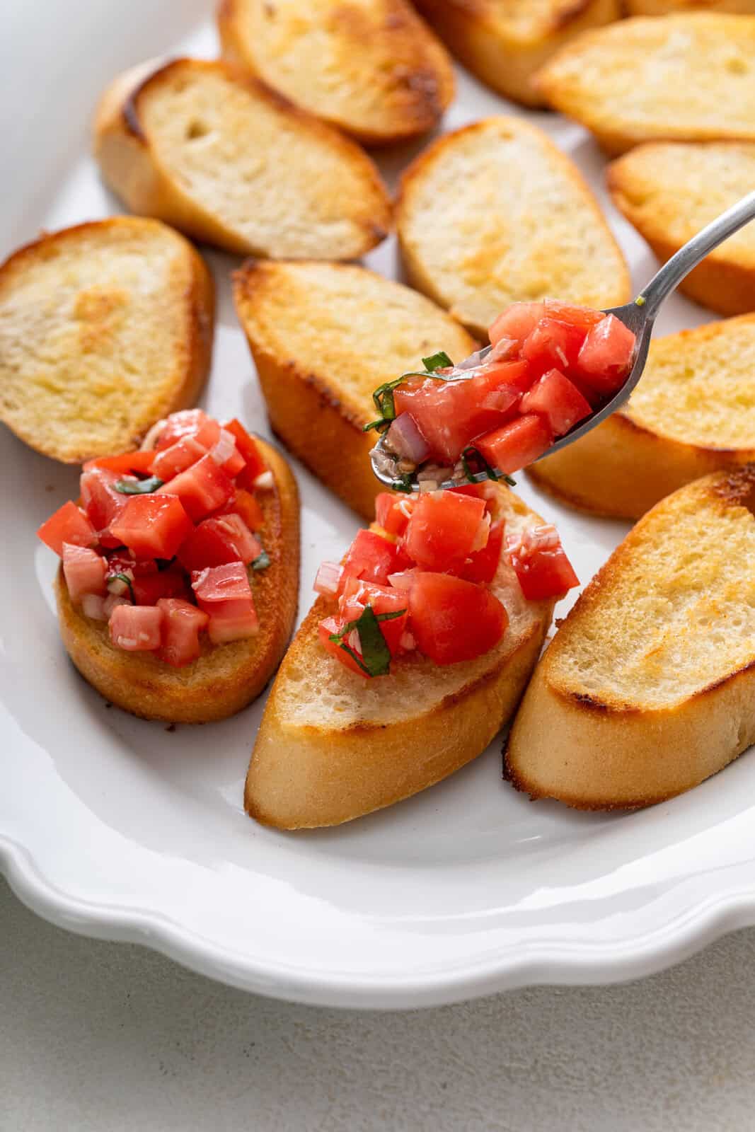 Spoon adding tomato topping to toasted baguette slices.