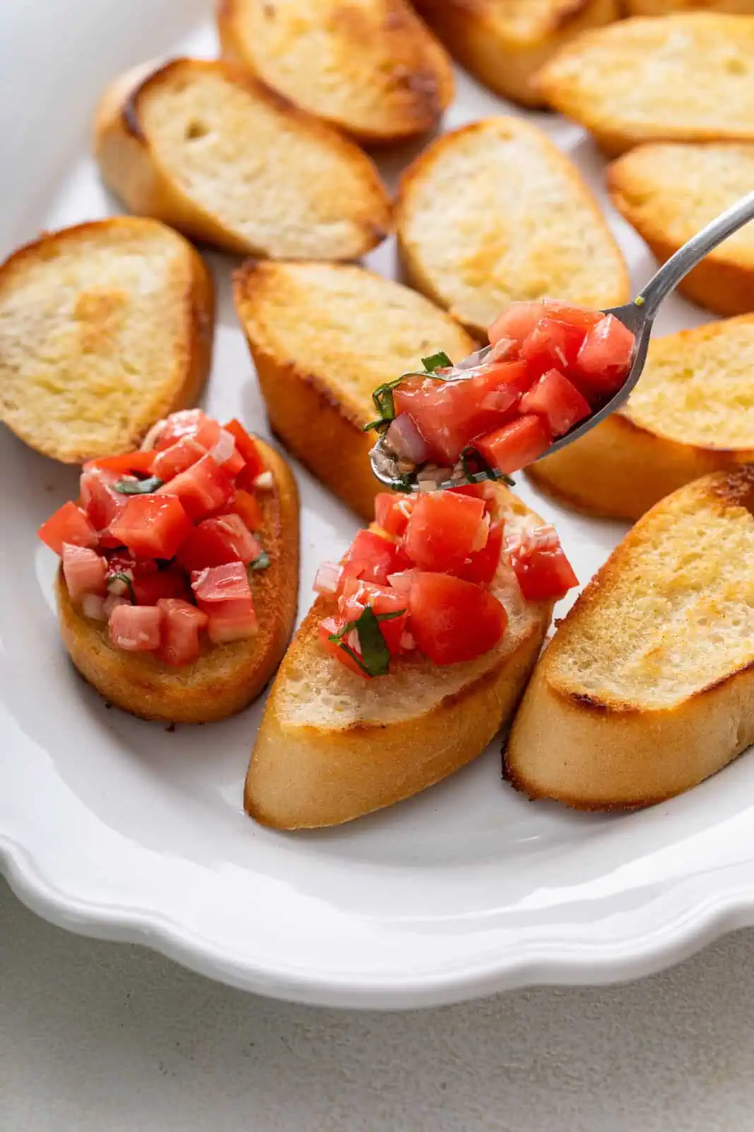 Spoon adding tomato topping to toasted baguette slices.