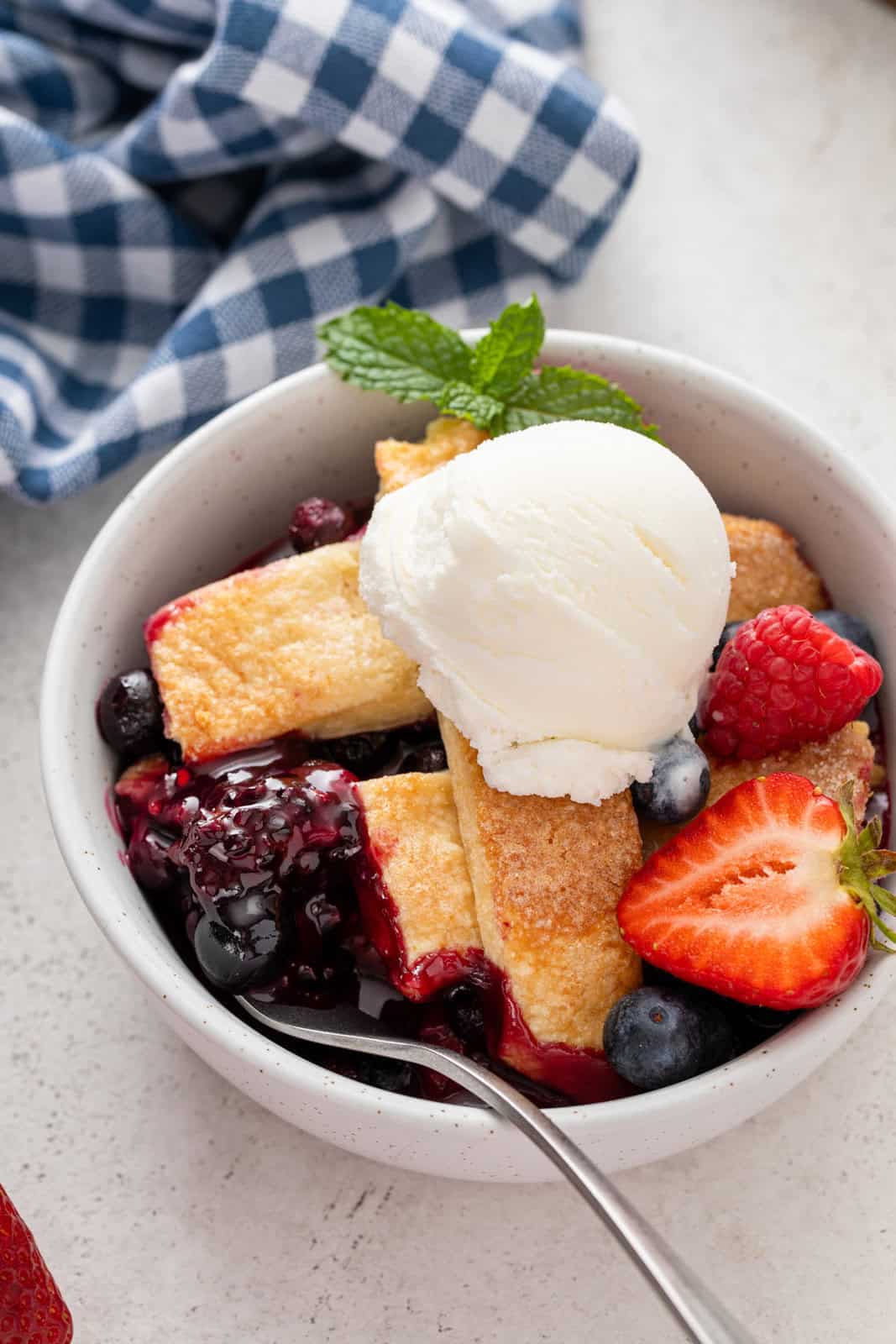 Spoon in a bowl of berry cobbler topped with a scoop of vanilla ice cream.