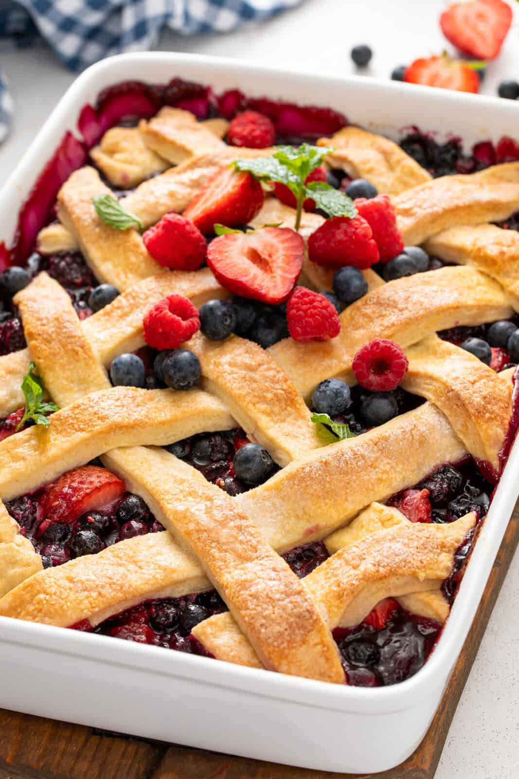 Lattice-topped berry cobbler in a white baking dish, garnished with fresh berries and fresh mint.