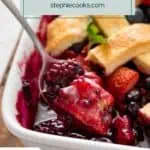 Spoon lifting a bite of mixed berry cobbler out of a baking dish. Text overlay includes recipe name.