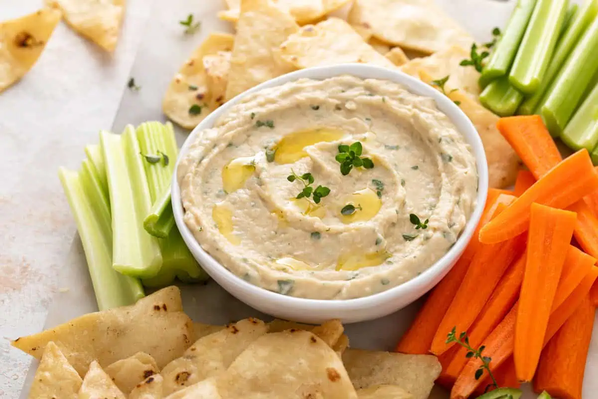 White bowl filled with garlic parmesan white bean dip surrounded by pita chips and fresh veggies.