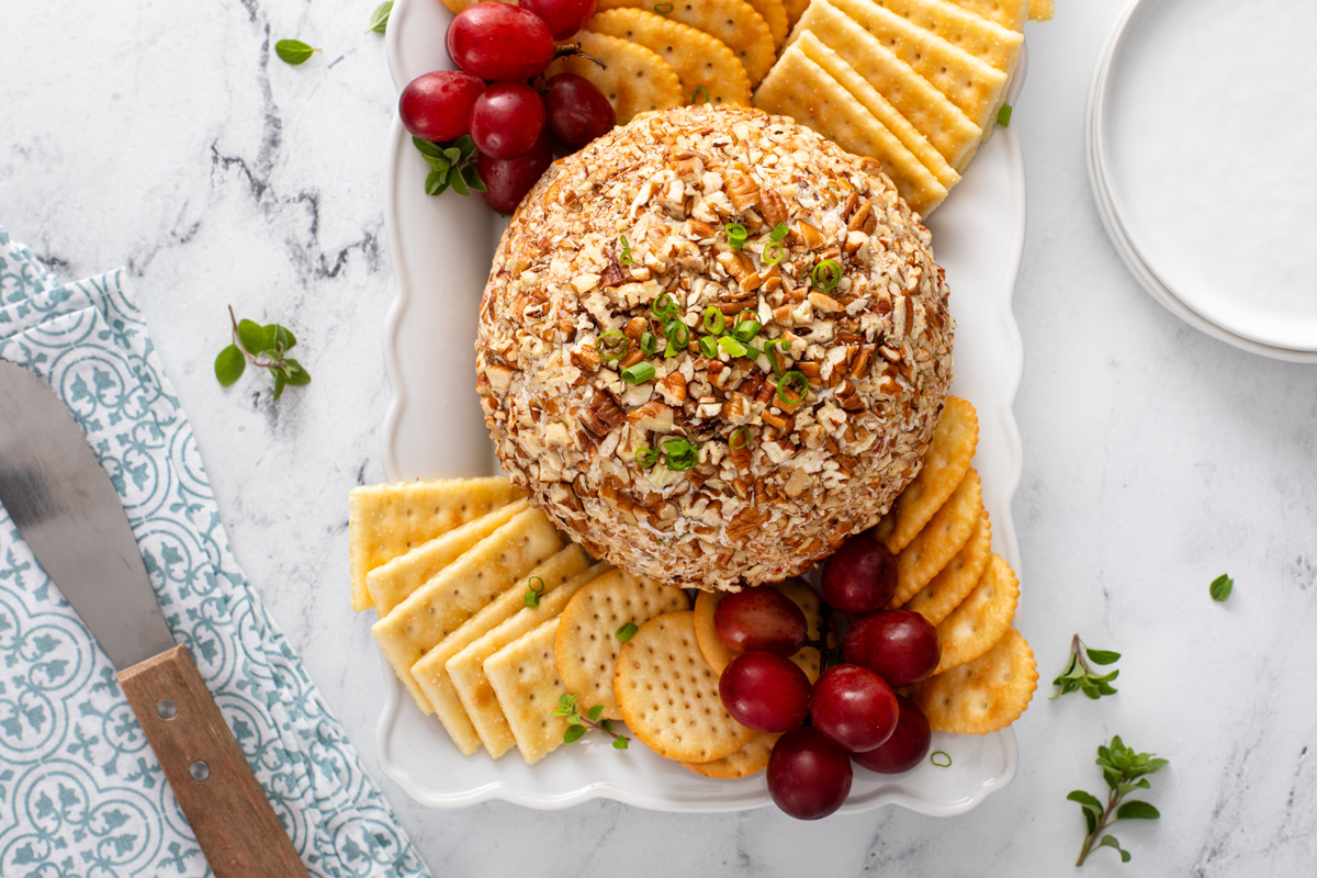 Overhead view of pineapple cheese ball on a white platter surrounded by crackers and grapes.