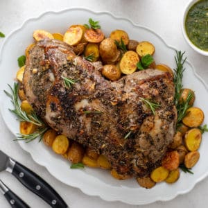 Roasted boneless leg of lamb on a white platter. Roasted potatoes surround the meat.